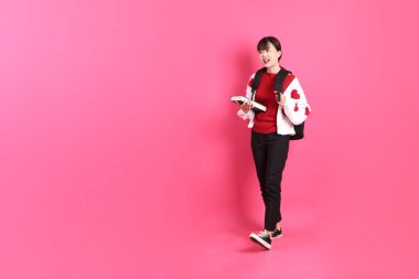 Kırmızı tişörtlü Asyalı kız öğrenci ya da günlük kıyafetler, kırmızı kalp şeklinde gömlek okul çantası ve kitabı pembe arka planda izole edilmiş yürüyüş jestleriyle taşıyor. Gen Z