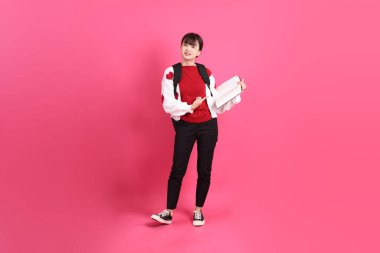 Kırmızı tişörtlü Asyalı kız öğrenci ya da günlük kıyafetler, kırmızı kalp şeklinde gömlek okul çantası ve kitap taşıyor pembe arka planda izole edilmiş bir işaret işareti var. Gen Z