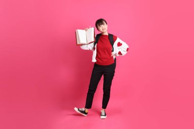 Kırmızı tişörtlü Asyalı kız öğrenci ya da günlük kıyafetler, kırmızı kalp şeklinde gömlek okul çantası ve kitap taşıyor ve pembe arka planda tek başına ayakta durup gülümsüyor. Gen Z