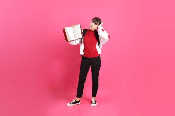 Kırmızı tişörtlü Asyalı kız öğrenci ya da günlük kıyafetler, kırmızı kalp şeklinde gömlek okul çantası ve kitabı pembe arka planda izole edilmiş düşünce jestleriyle taşıyor. Gen Z