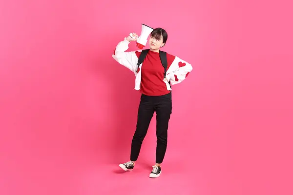Kırmızı tişörtlü Asyalı kız öğrenci ya da günlük kıyafetler, kırmızı kalp şeklinde gömlek okul çantası taşıyor ve pembe arka planda megafon taşıyor. Gen Z