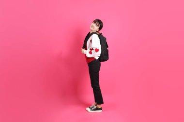 Kırmızı tişörtlü Asyalı kız öğrenci ya da günlük kıyafetler, kırmızı kalp şeklinde gömlek okul çantası taşıyor ve pembe arka planda sırıtıyor. Gen Z