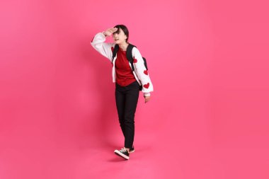 Kırmızı tişörtlü Asyalı kız öğrenci ya da günlük kıyafetler, kırmızı kalp şeklindeki gömlek okul çantasını taşır ve uzaklardan pembe arka planda izole bir şekilde bakar. Gen Z