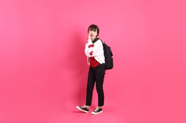 Kırmızı tişörtlü Asyalı kız öğrenci ya da günlük kıyafetler, kırmızı kalp şeklinde gömlek okul çantası taşıyor ve pembe arka planda izole edilmiş jestler yapıyor ya da fısıldıyor. Gen Z