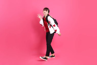 Kırmızı tişörtlü Asyalı kız öğrenci ya da günlük kıyafetler, kırmızı kalp şeklinde gömlek okul çantasını pembe arka planda izole edilmiş yürüyüş jesti ile taşıyor. Gen Z