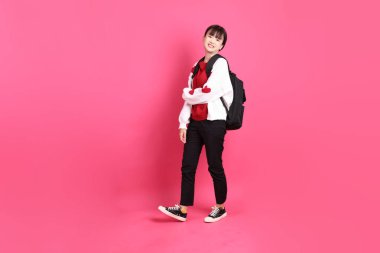 Kırmızı tişörtlü Asyalı kız öğrenci ya da günlük kıyafetler, kırmızı kalp şeklinde gömlek okul çantasını pembe arka planda izole edilmiş yürüyüş jesti ile taşıyor. Gen Z