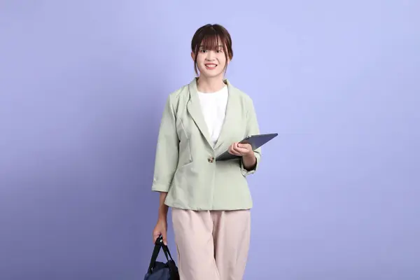 Resmi kıyafetli genç bir Asyalı iş kadını elinde tablet, ayakta duruyor ve gülümsüyor mor arka planda. Gen Z, staj, kariyer, ofis.