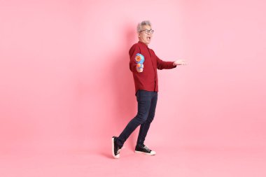 Mutlu SONGKRAN GÜNÜ. Resmi ve gündelik kırmızı tişörtlü Asyalı bir adam pembe arka planda izole edilmiş plastik su tabancası jesti yapıyor. Gen Z, Yaz, Tatil, Tayland Yeni Yıl Günü.