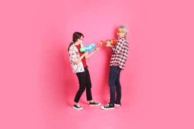 Mutlu SONGKRAN GÜNÜ. Asyalı son sınıf öğrencisi ve genç bayan, pembe arka planda izole edilmiş plastik su tabancası jesti ile Noel Baba şapkası ve günlük kırmızı kıyafetler giyerler. Gen Z, Yaz, Tatil, Tayland Yeni Yıl Günü.