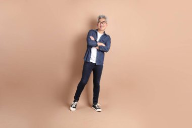 Asyalı bir üst sınıf öğrencisi ciddi bir jest yapmış, kolları çaprazlanmış ve sırıtarak kahverengi arka planda gezmiş, turist, şık, sıradan giyinmiş..