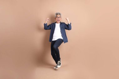 Asyalı son sınıf öğrencisi, evet jesti, heyecanlı, kahverengi arka planda izole edilmiş bir şekilde kutluyor. Turist, akıllı gündelik.