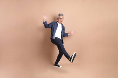 Kahverengi arka planda izole edilmiş eğlenceli ya da dans eden sıradan giyinmiş Asyalı son sınıf öğrencisi. dinamik duruş, akıllı gündelik.