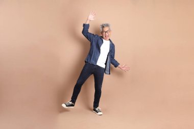 Kahverengi arka planda izole edilmiş eğlenceli ya da dans eden sıradan giyinmiş Asyalı son sınıf öğrencisi. dinamik duruş, akıllı gündelik.