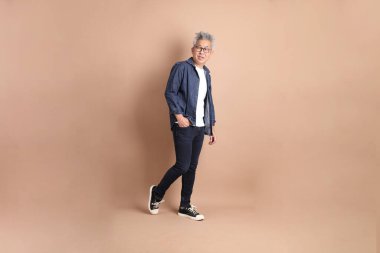 Kahverengi arka planda izole edilmiş yürüyüş jesti olan Asyalı son sınıf öğrencisi. Turist, akıllı gündelik.