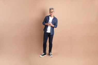 Kahverengi arka planda izole edilmiş Holding cep telefonu jesti ile Asyalı son sınıf öğrencisi. Turist, akıllı gündelik.