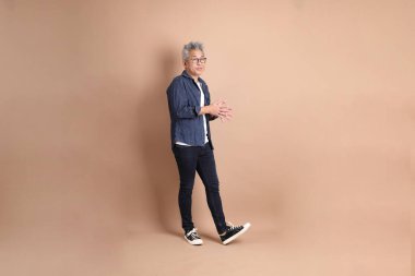 Kahverengi arka planda duran ve gülümseyen sıradan giyinmiş Asyalı son sınıf öğrencisi. Turist, akıllı gündelik.