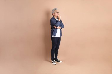 Kahverengi arka planda izole edilmiş bir düşünce jesti olan Asyalı son sınıf öğrencisi. Turist, akıllı gündelik.