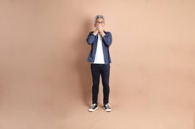 Kahverengi arka planda kapalı kalmış, ağzı kapalı, sıradan giyinmiş Asyalı bir adam. Turist, akıllı gündelik.