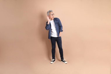 Kahverengi arka planda izole edilmiş dinleme ya da işitme jesti olan Asyalı son sınıf öğrencisi. Turist, akıllı gündelik.