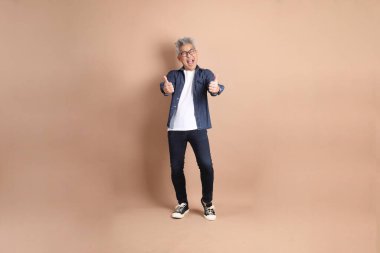 Asyalı son sınıf öğrencisi, sıradan giyinmiş, baş parmağı havada, ya da kahverengi arka planda izole edilmiş, tamam mı? Turist, akıllı gündelik.