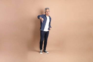 Kahverengi arka planda izole edilmiş baş parmak hareketleriyle sıradan giyinmiş Asyalı son sınıf öğrencisi. Turist, akıllı gündelik.