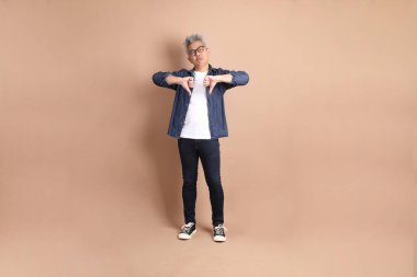 Kahverengi arka planda izole edilmiş baş parmak hareketleriyle sıradan giyinmiş Asyalı son sınıf öğrencisi. Turist, akıllı gündelik.