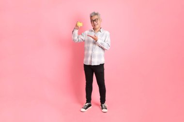 Resmi ve gündelik beyaz gömlekli Asyalı üst düzey bir adam. El hareketleriyle pembe arka planda izole edilmiş altın kredi kartı tutuyor..