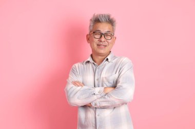 Asyalı son sınıf öğrencisi, resmi ve günlük beyaz gömlekli, ciddi bir jest, kolları çapraz ve pembe arka planda izole bir şekilde gülümseyen..