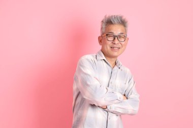 Asyalı son sınıf öğrencisi, resmi ve günlük beyaz gömlekli, ciddi bir jest, kolları çapraz ve pembe arka planda izole bir şekilde gülümseyen..