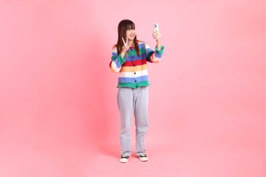 Renkli gökkuşağı kıyafetleri giymiş Asyalı bir kadın ya da selfie çekme jesti yaparak pembe arka planda izole edilmiş bir fotoğraf çekmek için fotoğraf makinesi tutan sıradan görünüşlü bir kadın. Gen Z