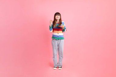 Renkli gökkuşağı kıyafetleri giyen Asyalı bir kadın ya da pembe arka planda izole edilmiş bir şekilde duran hediye kutusunu tutan sıradan görünüşlü bir kadın. Gen Z