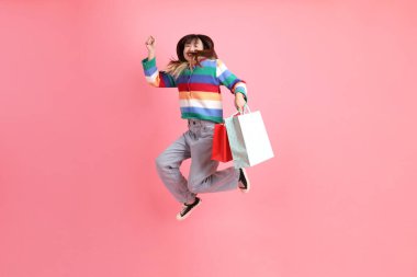 Renkli gökkuşağı kıyafetleri giyen Asyalı bir kadın ya da sıradan görünüşlü, pembe arka planda izole edilmiş alışveriş çantası taşıyan bir jest. Gen Z