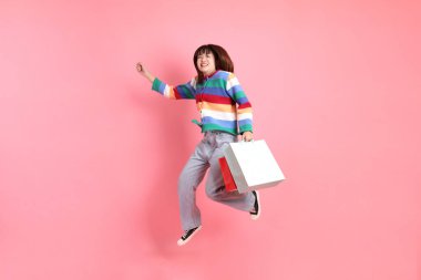 Renkli gökkuşağı kıyafetleri giyen Asyalı bir kadın ya da sıradan görünüşlü, pembe arka planda izole edilmiş alışveriş çantası taşıyan bir jest. Gen Z