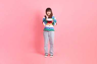 Renkli gökkuşağı kış kıyafetleri giyen Asyalı bir kadın ya da pembe arka planda saklanmış doğum günü pastası tutan bir jestle sıradan bir görünüm. Gen Z