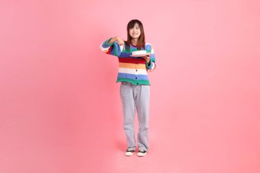 Renkli gökkuşağı kış kıyafetleri giyen Asyalı bir kadın ya da pembe arka planda saklanmış doğum günü pastası tutan bir jestle sıradan bir görünüm. Gen Z