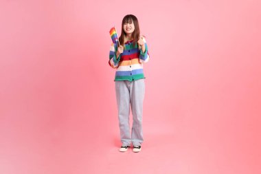 Renkli gökkuşağı kışı kıyafetleri giyen Asyalı bir kadın ya da başparmakları havada, pembe arka planda izole edilmiş, Gurur Bayraklarını Tutma jesti yapan sıradan görünüşlü bir kadın. Gen Z