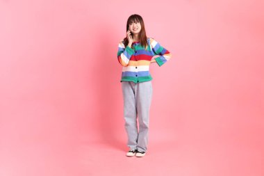 Renkli gökkuşağı kıyafetleri giyen Asyalı bir kadın ya da pembe arka planda izole edilmiş cep telefonu tutuş jesti içeren sıradan bir görünüm. Gen Z