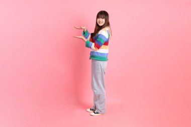 Renkli gökkuşağı kıyafetleri giyen Asyalı bir kadın ya da pembe arka planda izole edilmiş bir yeri işaret eden hareketli sıradan bir görünüm. Gen Z