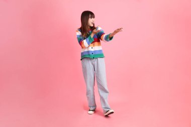 Renkli gökkuşağı kıyafetleri giyen Asyalı bir kadın ya da pembe arka planda izole edilmiş bir yeri işaret eden hareketli sıradan bir görünüm. Gen Z