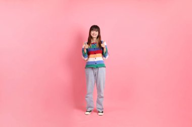 Renkli gökkuşağı kışı kıyafetleri giyen Asyalı bir kadın ya da pembe arka planda soyutlanmış bir jest yapan sıradan görünüşlü bir kadın. Gen Z