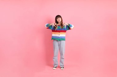 Renkli gökkuşağı kış kıyafetleri giyen Asyalı bir kadın ya da pembe arka planda izole edilmiş baş parmak hareketleriyle sıradan görünen bir kadın. Gen Z