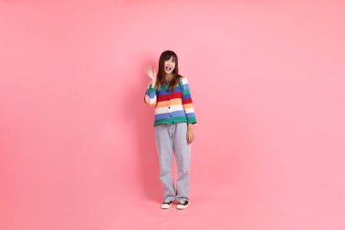 Renkli gökkuşağı kış kıyafetleri giyen Asyalı bir kadın ya da pembe arka planda izole edilmiş selamlama jesti içeren sıradan bir görünüm. Gen Z