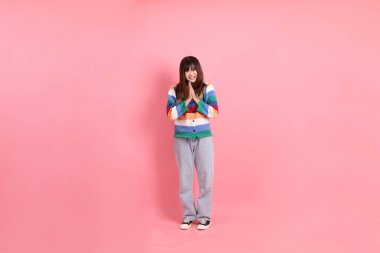 Renkli gökkuşağı kış kıyafetleri giyen Asyalı bir kadın ya da pembe arka planda izole edilmiş selamlama jesti içeren sıradan bir görünüm. Gen Z