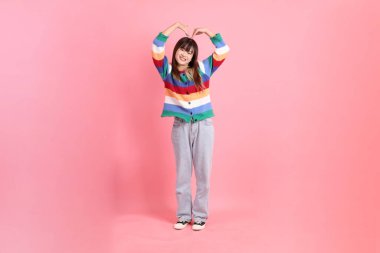 Renkli gökkuşağı kıyafetleri giyen Asyalı bir kadın ya da kalp şeklinde bir jest, küçük bir kalp, pembe arka planda izole edilmiş bir aşk. Gen Z