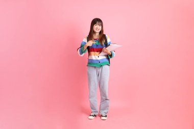 Renkli gökkuşağı kıyafetleri giyen Asyalı bir kadın ya da okul çantası ve kitap taşırken gelişigüzel görünen pembe arka planda izole edilmiş bir işaret hareketi yapan bir kadın. Gen Z