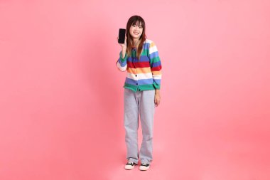 Renkli gökkuşağı kıyafetleri giyen Asyalı bir kadın ya da pembe arka planda izole edilmiş cep telefonu tutuş jesti içeren sıradan bir görünüm. Gen Z