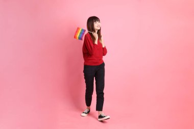 Pembe arka planda tek başına yürürken elinde Onur Bayrakları tutan kırmızı tişörtlü genç Asyalı kadın. Gen Z