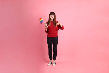 Pembe arka planda cep telefonunu kapalı tutarken elinde Gurur Bayrakları tutan kırmızı tişörtlü genç Asyalı kadın. Gen Z