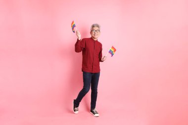 İki elinde de pembe arka planda izole edilmiş Rainbown Pride Bayrakları olan resmi ve sıradan kırmızı tişörtlü Asyalı kıdemli bir adam. LGBTQ + topluluğu