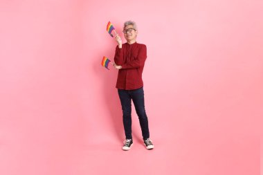 İki elinde de pembe arka planda izole edilmiş Rainbown Pride Bayrakları olan resmi ve sıradan kırmızı tişörtlü Asyalı kıdemli bir adam. LGBTQ + topluluğu